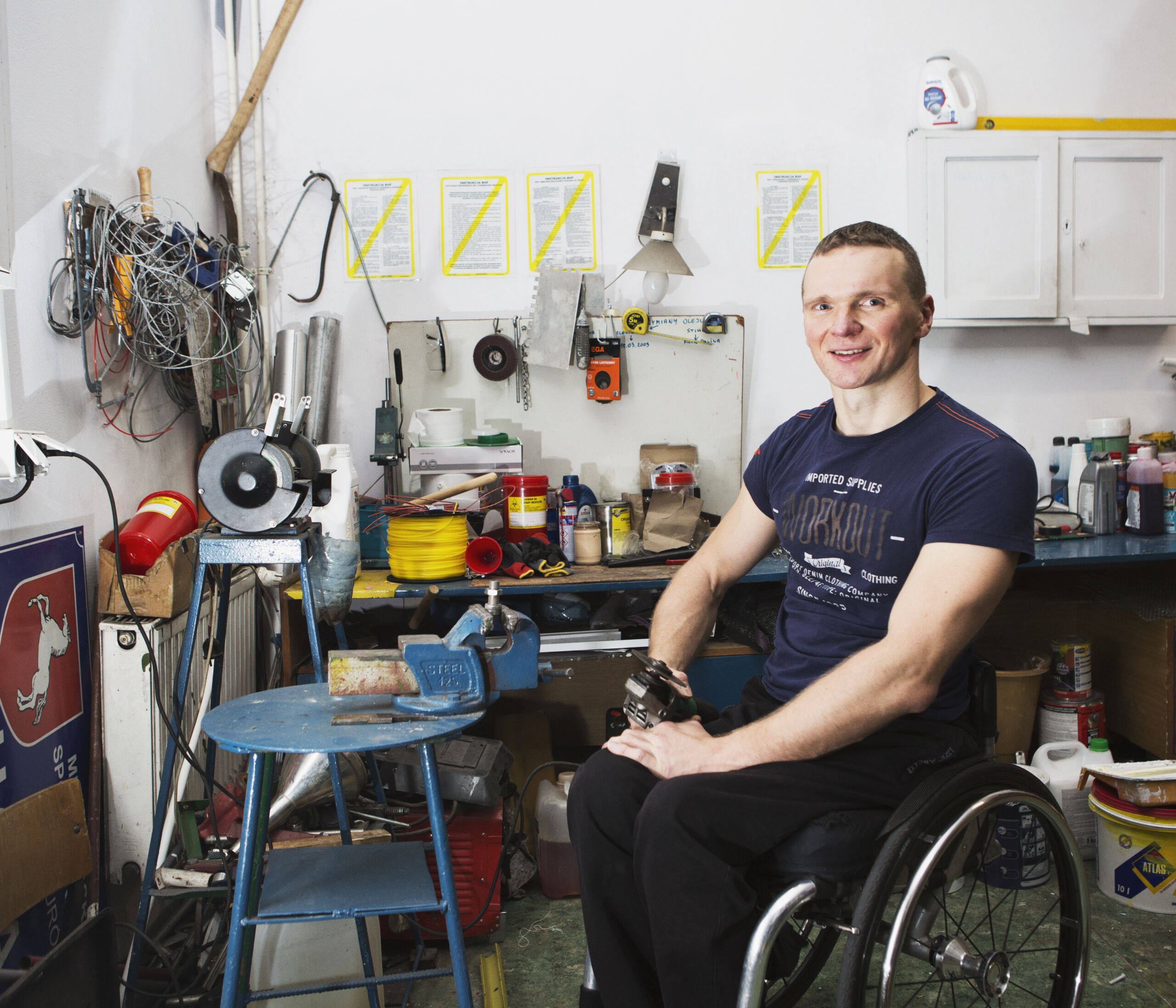 Mężczyzna siedzi na wózku inwalidzkim w warsztacie mechanicznym. W dłoni trzyma szlifierkę.